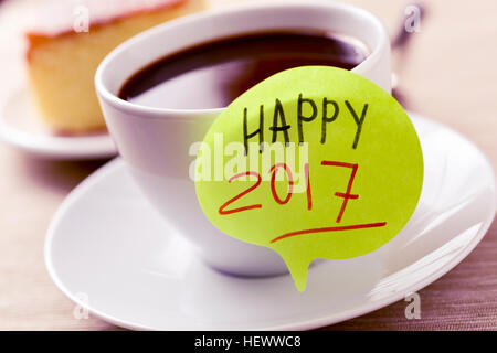 closeup of a yellow sticky note in the shape of a speech bubble with the text happy 2017 written in it attached to a white ceramic cup with coffee, on Stock Photo