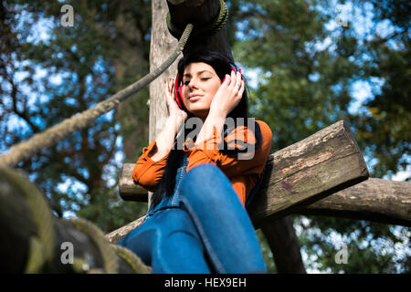 Young woman sitting on wooden climbing frame, wearing headphones Stock Photo