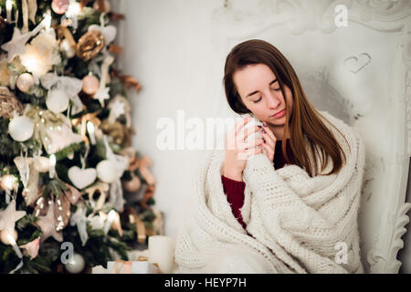 Young woman relaxing with a mug of coffee as she cuddles up in warm blanket on ancient commode. Her eyes closed and serene expression Stock Photo