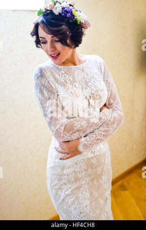 Beautiful bride in white lace dress and flowers in hair. Waiting in white room for her groom. Wedding morning. Stock Photo