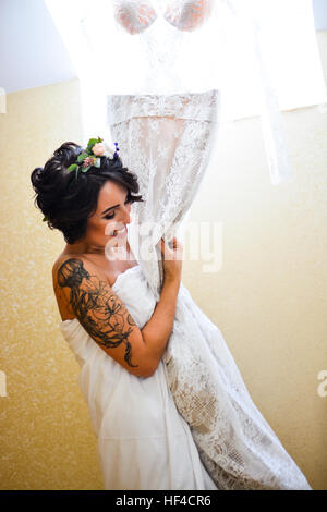 Beautiful smiling brunette bride posing with his wedding lace dress in hand and looking at mirror. Emotional girl wearing white  flowers in hair. Stock Photo