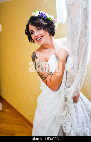 Beautiful smiling brunette bride posing with his wedding lace dress in hand and looking at mirror. Emotional girl wearing white  flowers in hair. Stock Photo