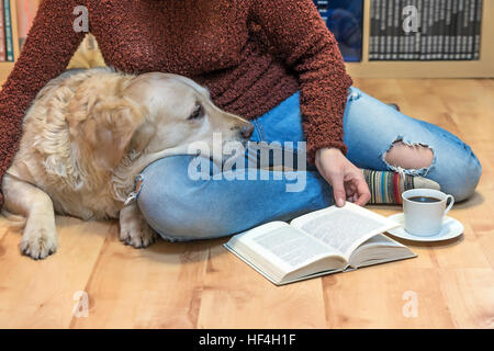 Woman is sitting down on the floor with Golden retriever dog. Open book and white cup of coffee are lying in front of them. Stock Photo