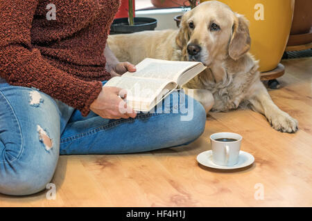 Woman is reading a book sitting down on the floor with Golden retriever dog. White cup of coffee is lying in front of her. Stock Photo