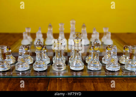 Glass Chess Pieces Set Up On Board In Starting Position Sitting On Wood Table With Yellow Wall Stock Photo