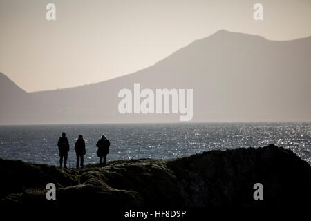 Three people looking towards the mainland coastal mountains on the Llyn Peninsula from Ynys Llanddwyn or Llanddwyn Island silhouetted against the setting sun, Anglesey Wales, UK