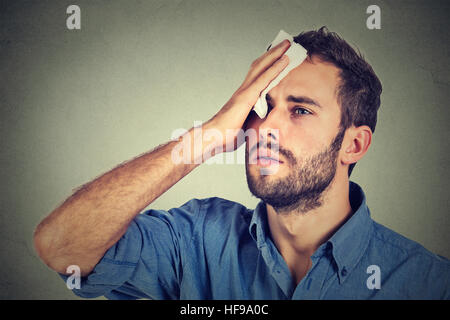 Tired man stressed sweating having fever headache isolated on gray wall background. Worried guy wipes sweat on his face Stock Photo