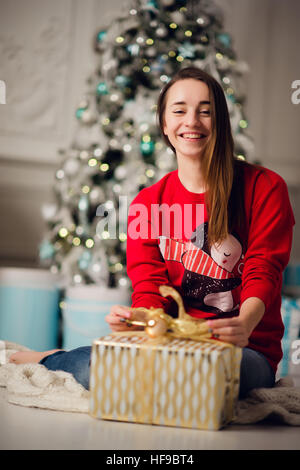 Portrait of beautiful woman with gift sitting by Christmas tree at home Stock Photo