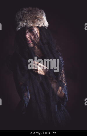 A slim petite young olive skinned woman girl wearing an old black lace wedding dress with a black lace veil over her face looking sad and lonely by herself alone Stock Photo