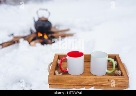 Romantic winter picnic. Two cups on a wooden tray in snow. Copper kettle over an open fire on background, blurred. Boiling kettle on firewood. Copy sp Stock Photo