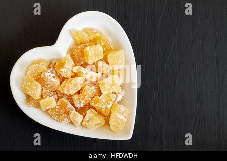 Candied ginger in heart shaped bowl on black wooden background. Top view with copy space Stock Photo