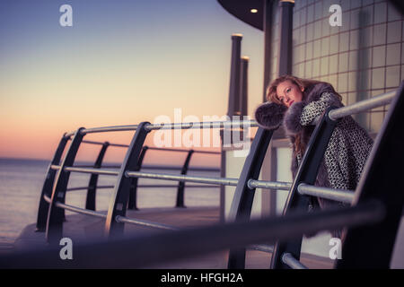 Photographic modeling as a career in the UK: A young slim attractive 20 year old caucasian woman girl with long blonde hair posing on seaside railings at dusk on a warm autumn evening, looking thoughtful and wistful, lonliness and sadness UK Stock Photo