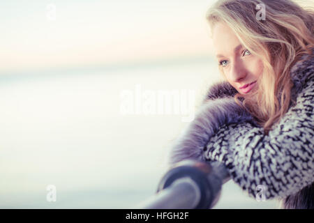 Photographic modeling as a career in the UK: A young slim attractive 20 year old caucasian woman girl with long blonde hair posing on seaside railings at dusk on a warm autumn evening, looking thoughtful and wistful, lonliness and sadness UK Stock Photo