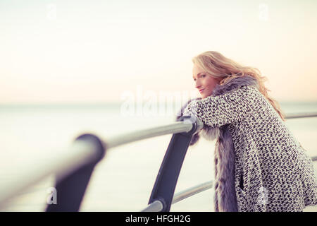 Photographic modeling as a career in the UK: A young slim attractive 20 year old caucasian woman girl with long blonde hair posing on seaside railings at dusk on a warm autumn evening, looking thoughtful and wistful, lonliness and sadness UK Stock Photo