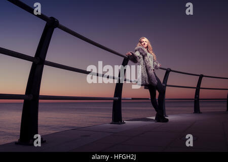 Photographic modeling as a career in the UK: A young slim attractive 20 year old caucasian woman girl with long blonde hair posing on seaside railings at dusk on a warm autumn evening UK Stock Photo