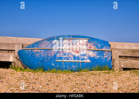 Old boat on its side against a wooden sea barrier on a shingle beach. Blue sky background. some sea grass around the boat Stock Photo