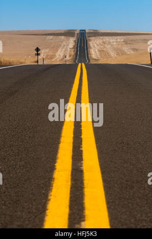 Bold yellow lines on a two lane blacktop highway running through golden wheat fields. Stock Photo