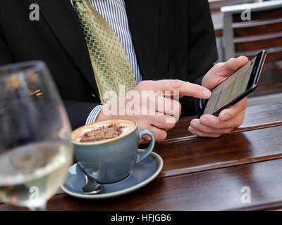 TEXTING CAFÉ BUSINESSMAN SMART Close-up on mature businessman's hands texting with his iPhone 7 plus smartphone sitting at alfresco restaurant situation with glass of white wine and cappuccino coffee Stock Photo