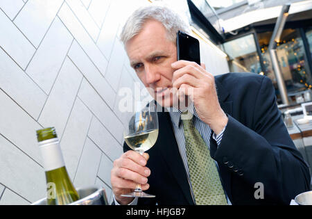 Mature city executive businessman, looking pensive listening seriously on his iPhone 7 plus mobile telephone at alfresco café restaurant table holding glass of white wine with wine bottle and chiller in foreground Stock Photo