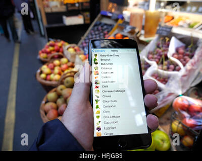 IPHONE SHOPPING LIST FRUIT & VEG Man holding black smartphone iPhone , with shopping list suggestions on its interactive screen, fruit and veg produce market stall behind. Borough Market London UK Stock Photo