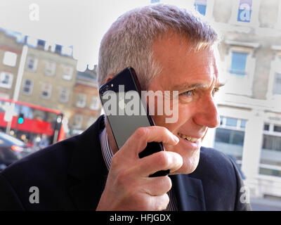 Smiling city businessman sitting at outdoor café talking on his jet black smartphone iPhone 7 plus with typical busy London street background Stock Photo