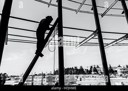 Silhouetted industrial shot of construction worker who is using the wooden ladder to welding metal frame. Stock Photo