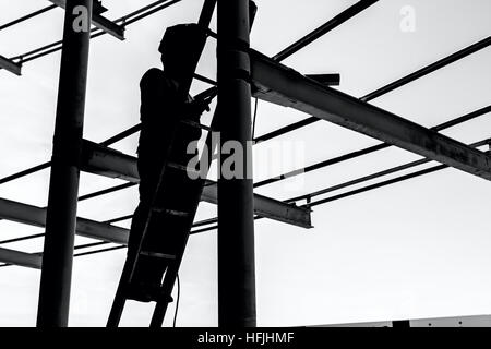 Silhouetted industrial shot of construction worker who is using the wooden ladder to welding metal frame. Stock Photo