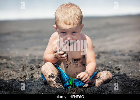 Funny happy baby boy with dirty body and sly face playing game, eating black sand. Family travel lifestyle, holiday recreation