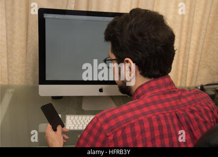 Young man sitting in front of a desktop computer with a smartphone Stock Photo