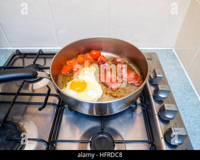 Bacon egg and tomatoes being fried in sunflower oil in stainless steel frying pan on a gas hob Stock Photo