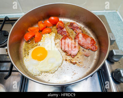 Bacon egg and tomatoes being fried in sunflower oil in stainless steel frying pan on a gas hob Stock Photo