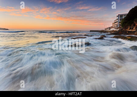 Bondi beach surfing exerience at sunrise with streaming wave on shore boulders in view of public rock pull and icebergs cafe. Stock Photo