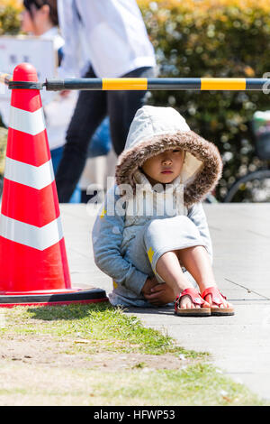 Japan, Kumamoto. Little child, Japanese girl sitting in bright sunshine with oversized coat and hood on, hands clasped under legs. Facing Stock Photo
