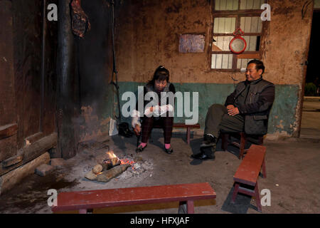 A woman stirs the fire with tongs while her husband looks on as they sit in the living room of their farmhouse near the open fire to keep warm in wint Stock Photo