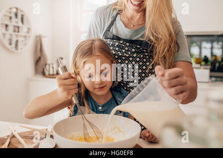 Cute little girl and her mother mixing batter in the bowl. Mother pouring milk with daughter whisking the batter. Stock Photo