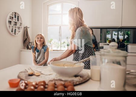 Happy little girl standing in kitchen and her mother cooking food. Mother and happy daughter baking in kitchen.