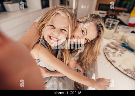 Smiling little girl and woman in kitchen taking selfie. Happy young mother and daughter cooking food. Stock Photo