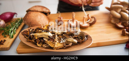In the foreground, a wooden dish holds dried up mushrooms. In the background, a woman's elegant hands are stringing fresh mushrooms together as she is Stock Photo