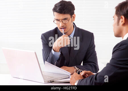 Young professional man in deep thought holding pen between his teeth looking at laptop computer while discussing work with another professional man Stock Photo