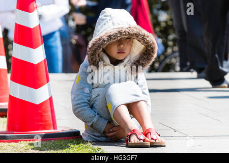 Japan, Kumamoto. Little child, Japanese girl sitting in bright sunshine with oversized coat and hood on, hands clasped under legs. Facing