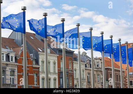 A line of EU flags flying on metal poles in front of the Berlaymont Building in Brussels with traditional houses in the background.