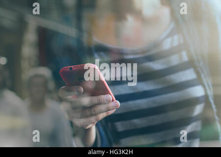 Young Caucasian woman with stripped blouse using a phone with a pink cover seen through a cafe window with people reflected Stock Photo