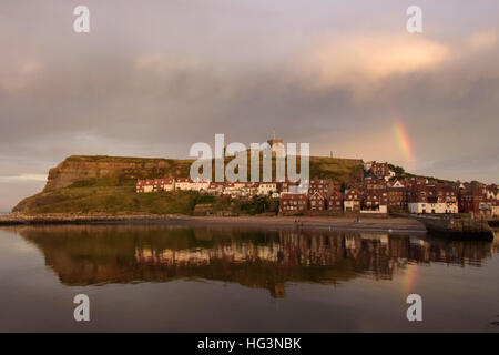 View of the East Cliff (houses, St. Mary's Church, abbey ruins and rainbow) reflected in the sea - Whitby, North Yorkshire, GB. Stock Photo