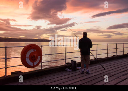 Scenic Whitby & silhouetted against dramatic, bright colourful sunset sky, man (angler) is sea fishing from West Pier - North Yorkshire, England, UK. Stock Photo