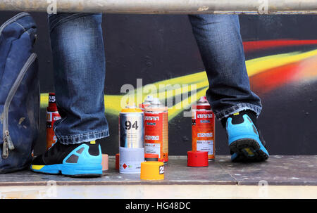 The legs of a graffiti artist standing on a platform whilst working on a piece of art. Cans of spray paint are at his feet Stock Photo
