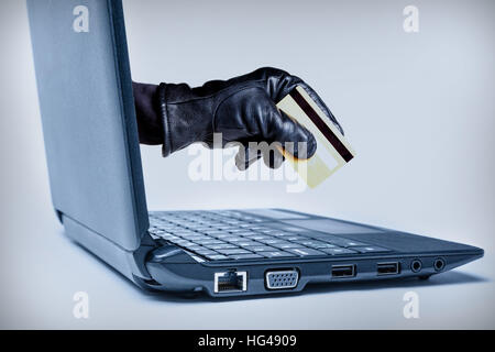 A gloved hand reaching out through a laptop holding debit or credit card, signifying a cybercrime or Internet theft. Stock Photo