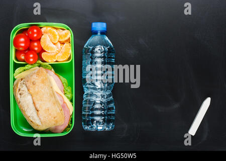 Sandwich, small tomatoes, tangerine in plastic lunch box and bottle of water on black chalkboard. Stock Photo