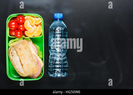 Sandwich, small tomatoes, tangerine in plastic lunch box and bottle of water on black chalkboard. Stock Photo