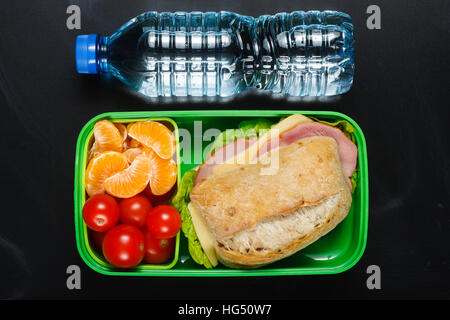 Sandwich, small tomatoes, tangerines in plastic lunch box and bottle of water on black chalkboard. Stock Photo