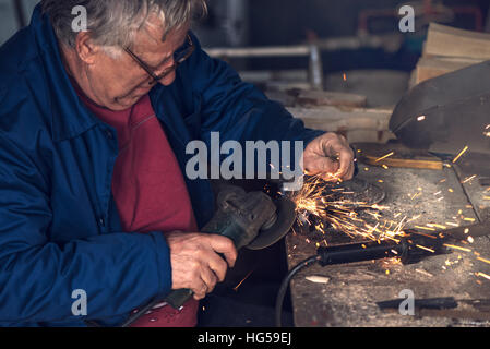Mature male worker grinding piece of metal with grinder tool in workshop without protective equipment Stock Photo